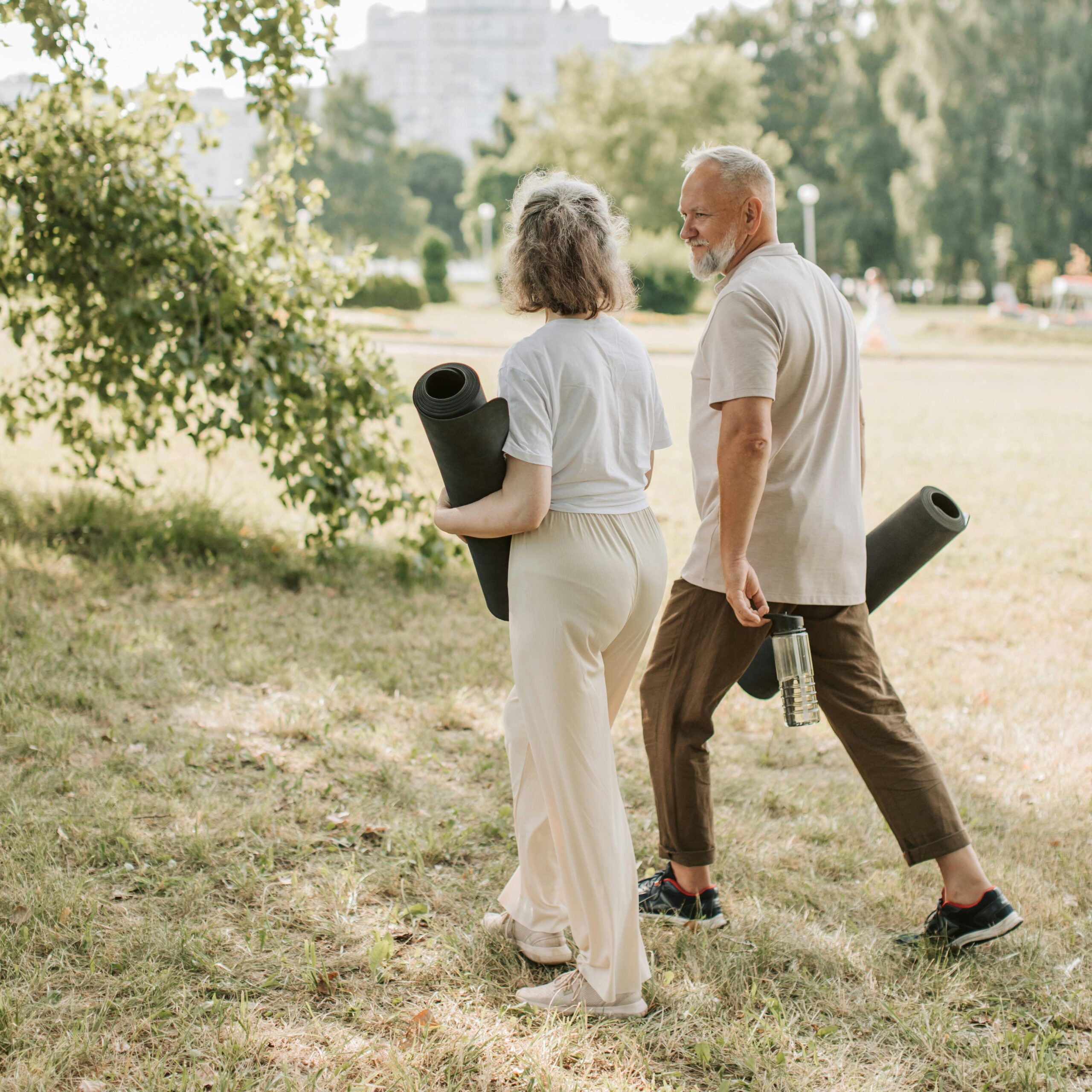 Two older adults walk on grass in a park, each carrying a rolled yoga mat. Embracing active aging, one also holds a water bottle. Trees and buildings are visible in the background.