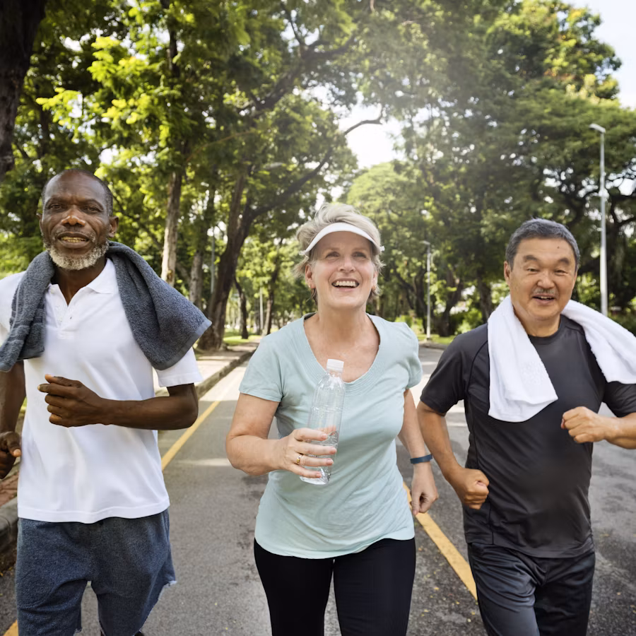 Three older adults, two men and one woman, jog together in a park on a sunny day, smiling and wearing athletic clothing with towels around their necks—embracing longevity and healthy aging through an active lifestyle.